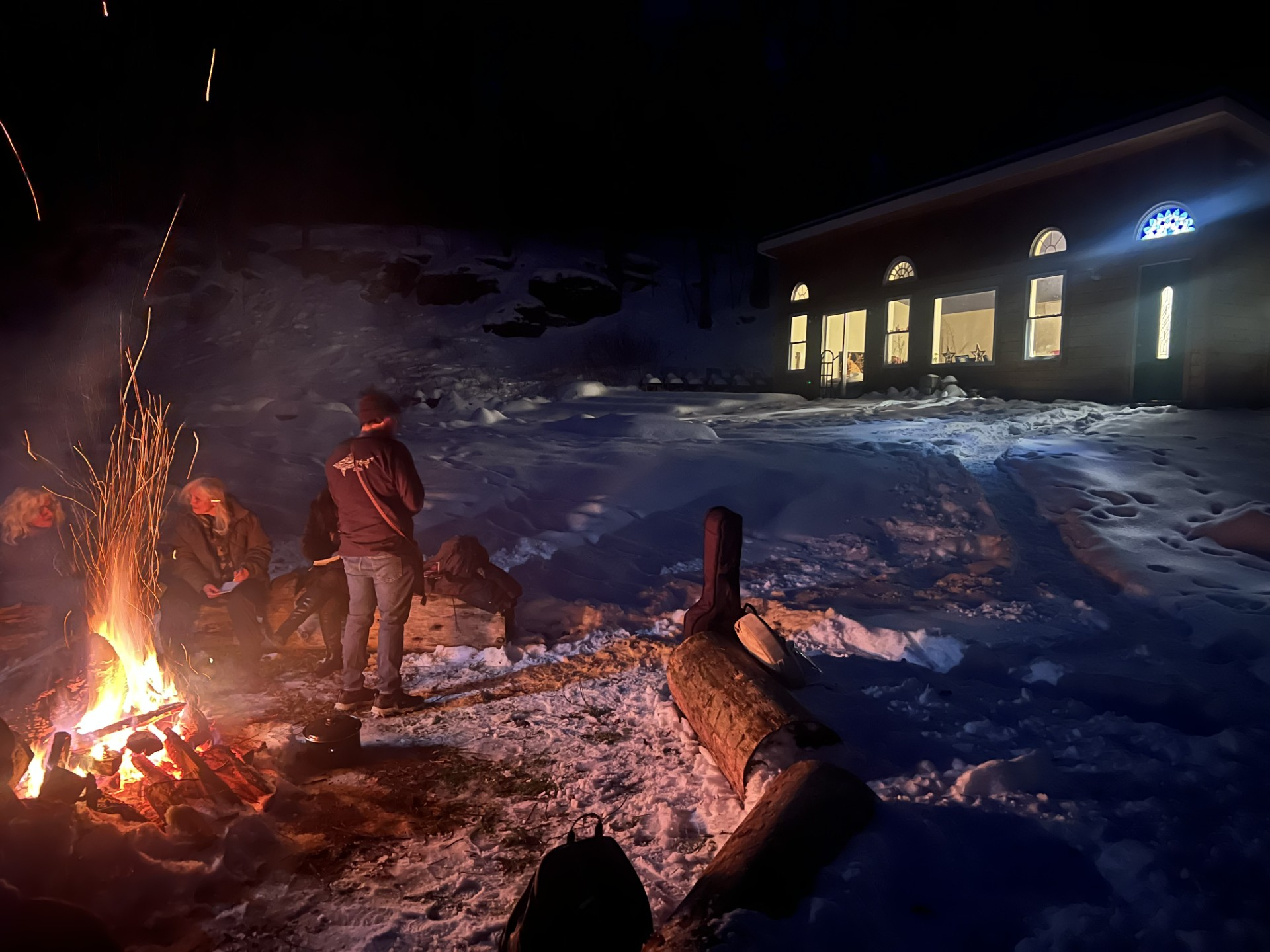 Guests gathered around a bonfire in the snow outside the lit studio at night